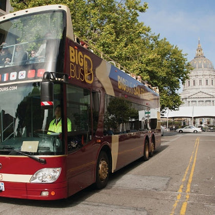 Double-decker tour bus labeled "Big Bus" on a city street with people on the upper deck, near a domed building and trees.
