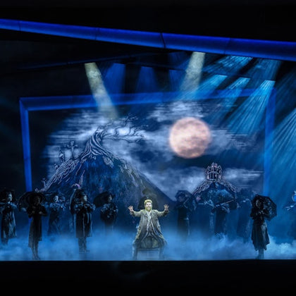 A theatrical performance with a moonlit backdrop, fog effects, and actors in dark costumes on stage under dramatic lighting.
