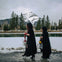 Two people in dark cloaks walk by a snowy river, holding lanterns. Snowy mountain and evergreen trees in the background.