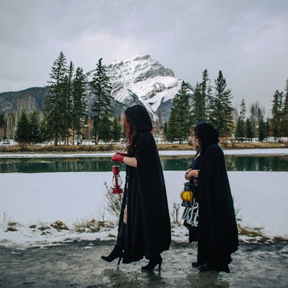 Two people in dark cloaks walk by a snowy river, holding lanterns. Snowy mountain and evergreen trees in the background.