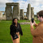 A person smiles while posing for a photo near Stonehenge, with another person holding a smartphone to take the picture.