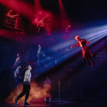 A dramatic stage performance with a person flying under spotlight, surrounded by actors and red lighting effects.