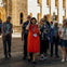 A group of people wearing lanyards and earbuds, walking near a historic stone building with arched windows and doorways.