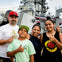 A smiling family of four poses in front of a large naval ship with patriotic decorations, giving thumbs-up gestures.