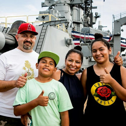A smiling family of four poses in front of a large naval ship with patriotic decorations, giving thumbs-up gestures.
