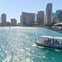 A water taxi is cruising on a blue waterway with a city skyline of tall buildings in the background under a clear sky.