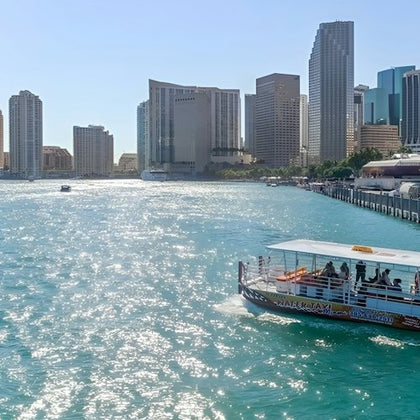 A water taxi is cruising on a blue waterway with a city skyline of tall buildings in the background under a clear sky.