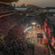 A large outdoor event with a crowd in a stadium, a lit stage with performers, and a city skyline in the background at dusk.