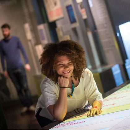 Smiling person using an interactive exhibit in a museum, with another person in the background.