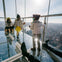 Two children and an adult enjoy a glass floor observation deck high above a cityscape on a sunny day.