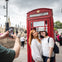 Two people pose in front of a red telephone booth in a city as another person takes their photo with a smartphone.