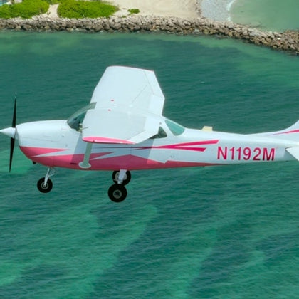 Small pink and white airplane flying over clear green ocean water near a rocky shoreline.