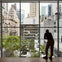 A person observes a cityscape through a large window, overlooking a garden courtyard with surrounding buildings.