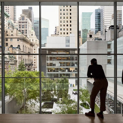 A person stands indoors, looking out at a cityscape and garden through large glass windows.