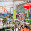 Indoor amusement park with colorful rides, roller coasters, and a crowd of visitors under a high glass ceiling.