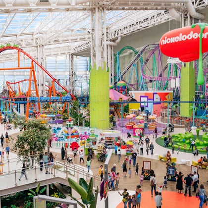 Indoor amusement park with colorful rides, roller coasters, and a crowd of visitors under a high glass ceiling.