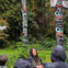 A group of people stand near totem poles in a forested area, with a guide in the foreground speaking.