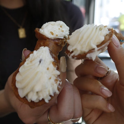 Two hands holding cannoli filled with cream, partially broken open, against a blurred background.