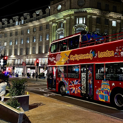 A red double-decker sightseeing bus with colorful designs parked on a nighttime street in front of a lit-up building.