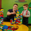 A man and a boy build a colorful structure with Lego bricks on a yellow table against a green background.