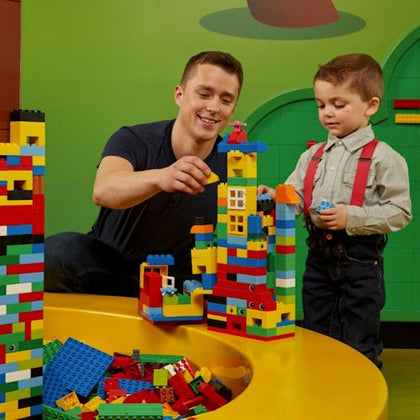 A man and a boy build a colorful structure with Lego bricks on a yellow table against a green background.