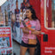 A woman in a pink shirt holding brochures stands near a red bus with promotional signs and posters in the background.