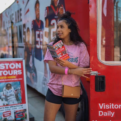 A woman in a pink shirt holding brochures stands near a red bus with promotional signs and posters in the background.