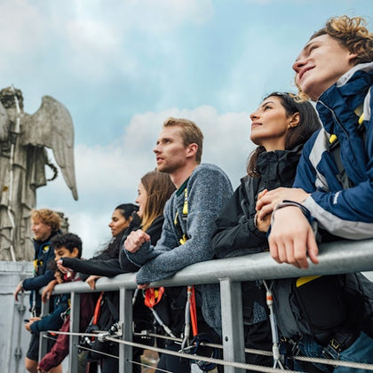 A group of people stands together on a platform, wearing harnesses, looking outward. A stone angel statue is visible nearby.