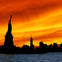 Silhouette of the Statue of Liberty and New York City skyline at sunset with a vivid orange sky over water.
