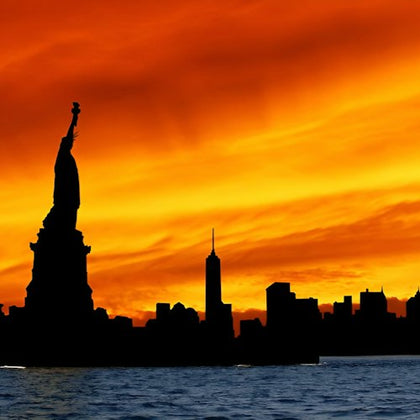 Silhouette of the Statue of Liberty and New York City skyline at sunset with a vivid orange sky over water.
