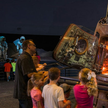 A group of children and an adult observe a spacecraft interior on display. Astronaut mannequins are visible in the background.