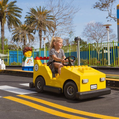 A child driving a yellow Lego-themed car on a track, with a Lego figure and palm trees in the background.