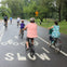 People ride bicycles on a paved pathway in a park, with large trees and greenery around. The road has "SLOW" painted on it.