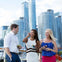 Three people standing outdoors, smiling, and holding drinks, with modern high-rise buildings in the background.