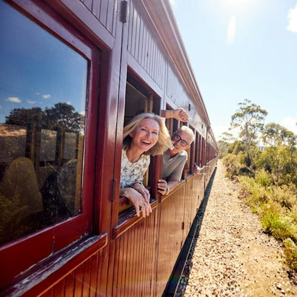 Two people smiling and leaning out of the windows of a red vintage train traveling through a sunny countryside.