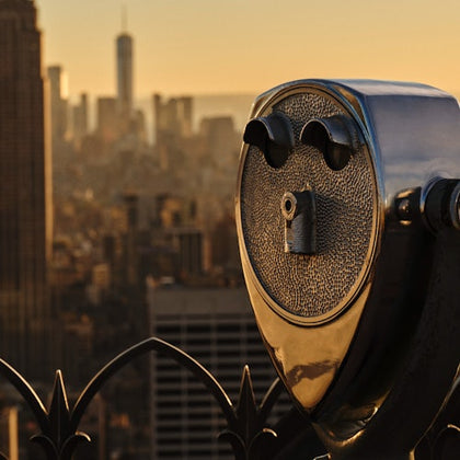 A coin-operated binocular viewer overlooks a cityscape at sunset, with buildings and a soft orange sky in the background.