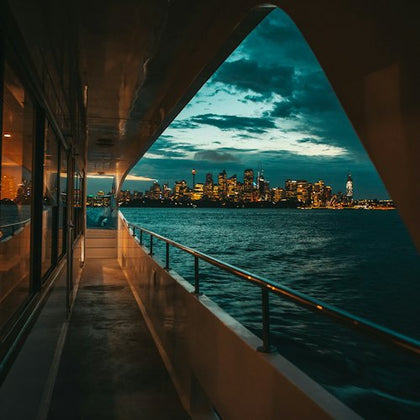 View from a boat of a city skyline at dusk. Water reflects city lights; dramatic clouds enhance the sky.