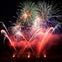 Colorful fireworks burst in various shapes over a dark night sky, with red and white trails illuminating the smoke below.