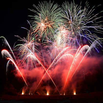 Colorful fireworks burst in various shapes over a dark night sky, with red and white trails illuminating the smoke below.