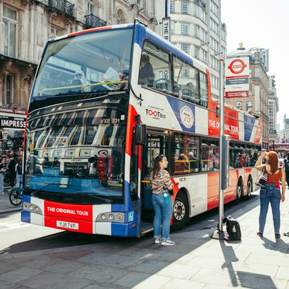 A double-decker tour bus parked at a bus stop in an urban setting with people nearby, surrounded by historic buildings.