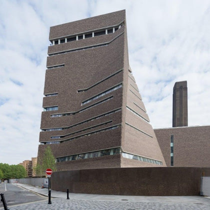 A modern, angular brick building with slanted walls and narrow windows, set against a partly cloudy sky.