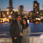 A couple smiling on a boat with a city skyline and blurred lights in the background at dusk.