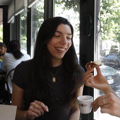 A woman smiling indoors near a large window, holding a small object while another hand offers a pastry and a cup of coffee.
