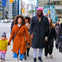 A group of people walking on a city sidewalk on a bright day; the central individuals are wearing colorful coats.