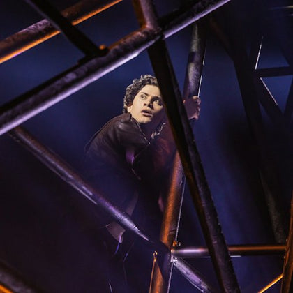 A person with curly hair clings to a metal structure, illuminated by dramatic lighting, with a dark and smoky background.