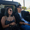 Two people sitting in the back of a vehicle, smiling, wearing sunglasses, with a sunny outdoor setting in the background.