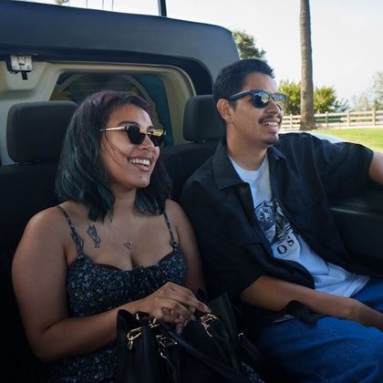Two people sitting in the back of a vehicle, smiling, wearing sunglasses, with a sunny outdoor setting in the background.