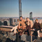 Four people sitting on a high steel beam overlooking a cityscape, with tall buildings visible in the background.
