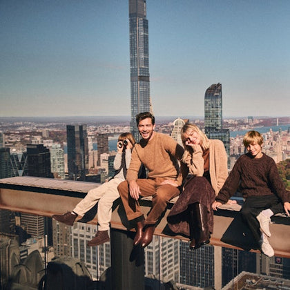 Four people sitting on a high steel beam overlooking a cityscape, with tall buildings visible in the background.