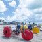 Beach scene with oversized tricycles featuring large red and yellow wheels on the sand, under a partly cloudy sky.